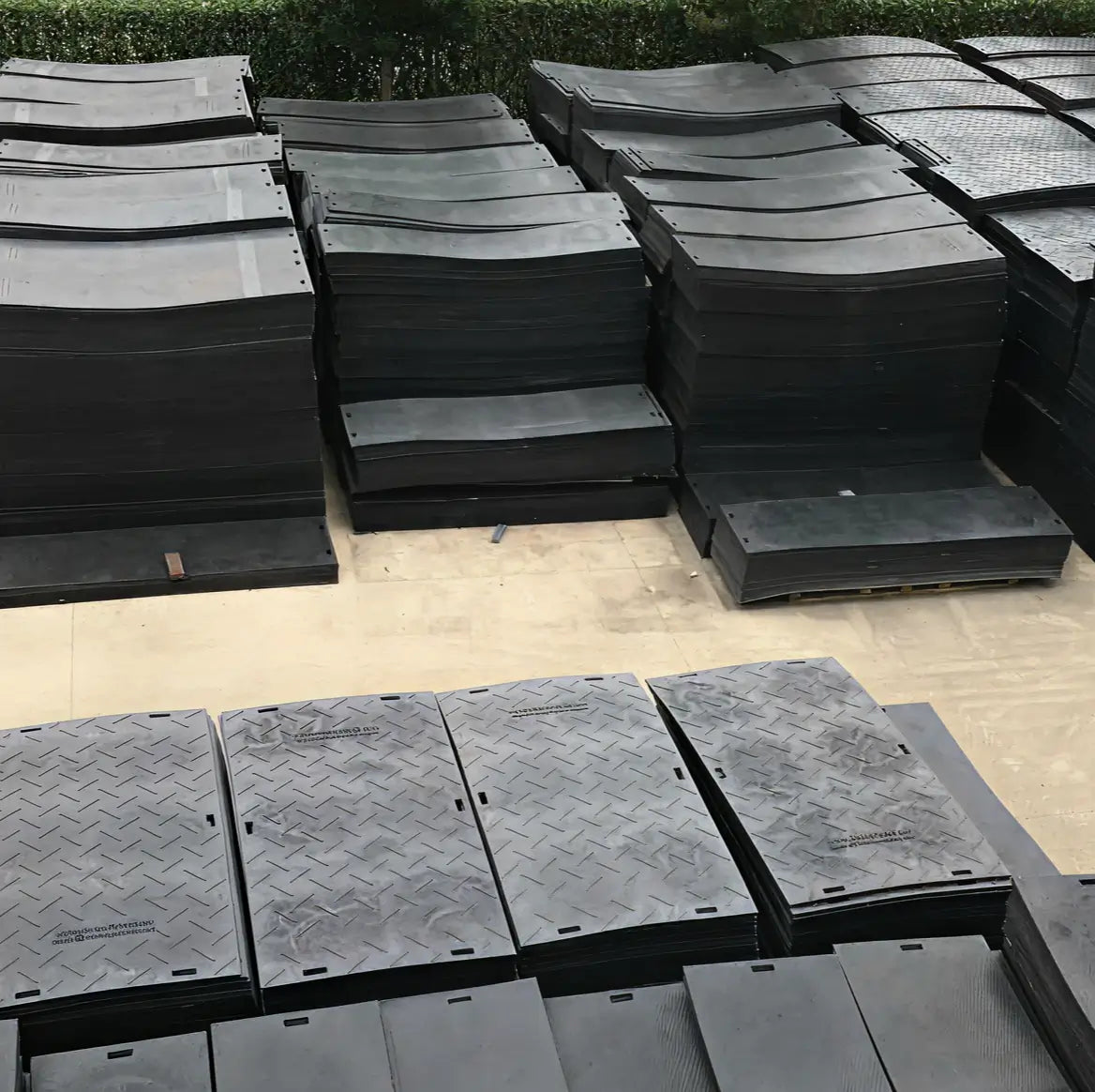 Stacks of black heavy-duty ground protection mats with diamond-tread surfaces stored outdoors on pallets
