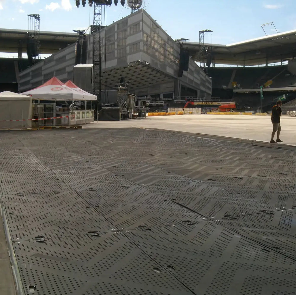 Temporary perforated ground protection mats covering a stadium floor during event setup, with lighting rigs and stage scaffolding visible in the background