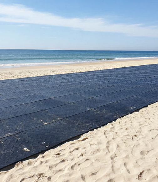Wide black beach access mats laid flat on sandy shore near the water's edge.