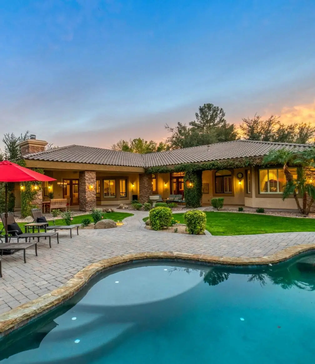 Luxury backyard view of a southwestern-style home at sunset featuring a curved swimming pool, stone patio, lounge chairs, and a red umbrella.