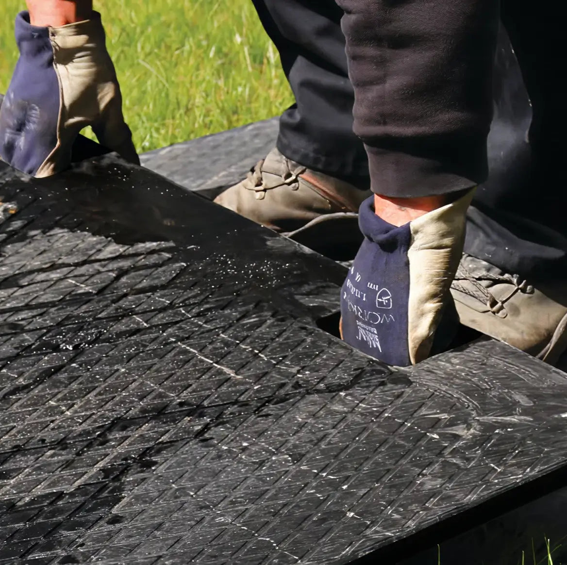 Close-up of a worker wearing gloves connecting two interlocking black ground protection mats on grass

